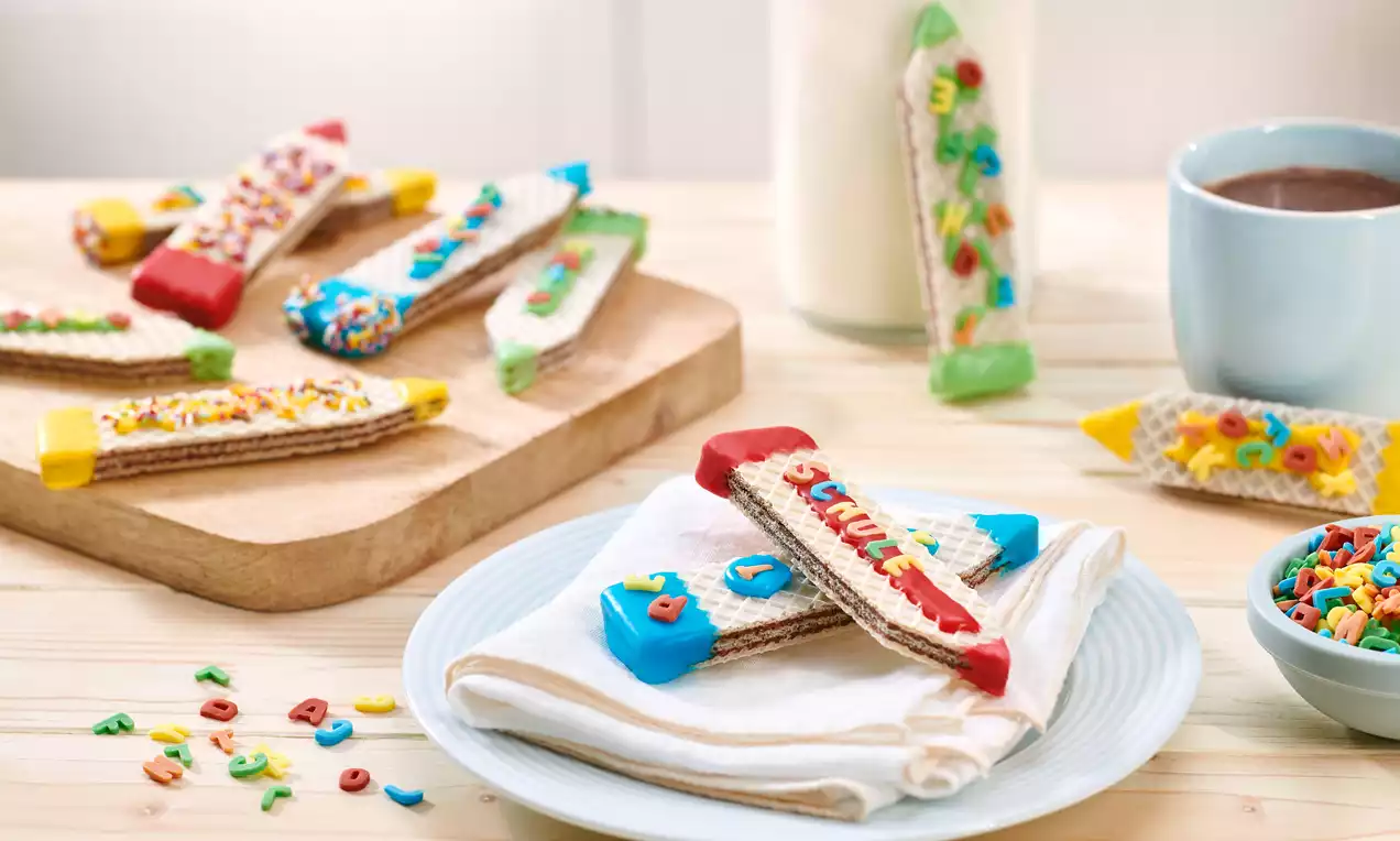 Bunte Waffel-Stifte mit knuspriger Textur, farbigem Zuckerguss und Dekor, teils auf Holzbrett, teils auf Teller mit Serviette angerichtet