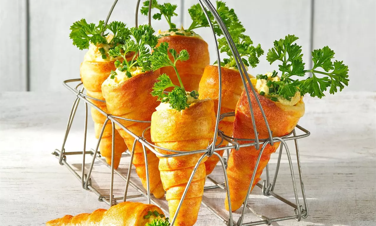 Zlatisté pečené mrkvičky kónického tvaru plnené krémovou náplňou, ozdobené zelenou petržlenovou vňaťou, uložené v kovovom koši