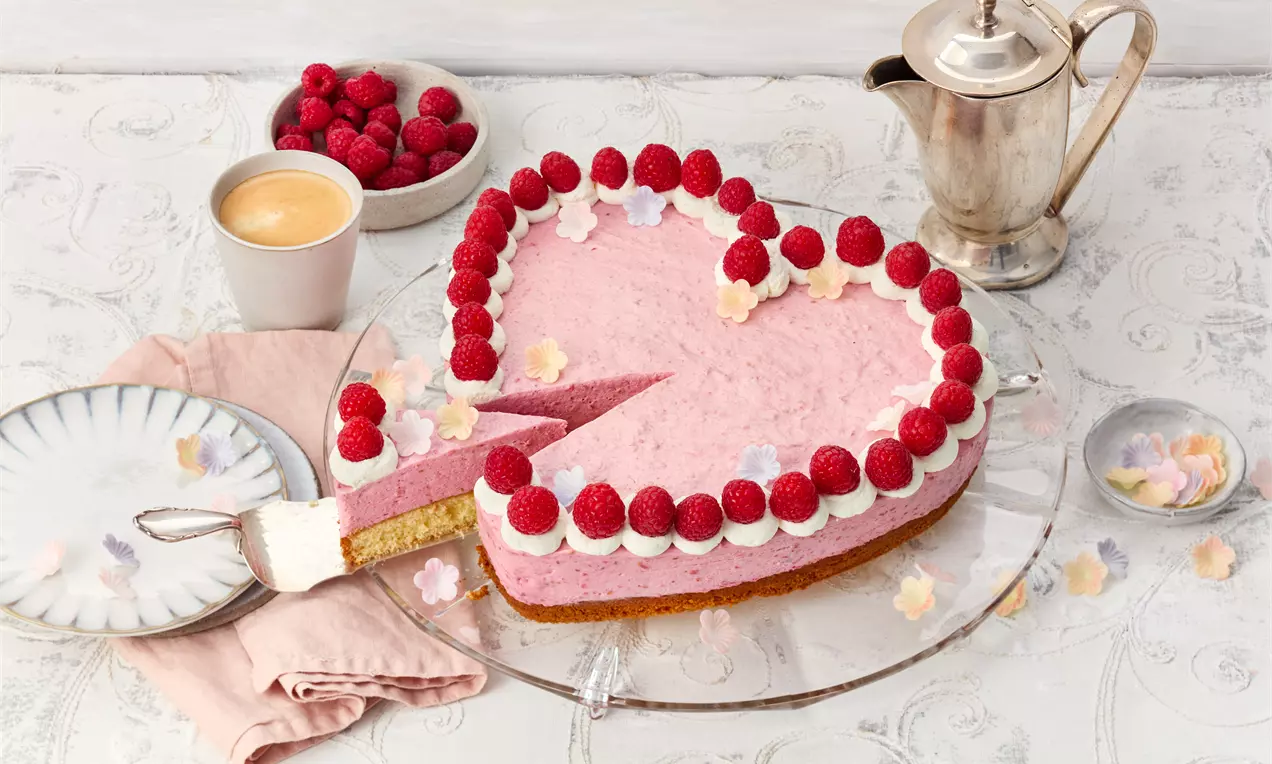 Herzförmige Torte mit rosa Creme, dekoriert mit weißen Sahnetupfen und roten Himbeeren auf einer Glasplatte, serviert mit Kaffee
