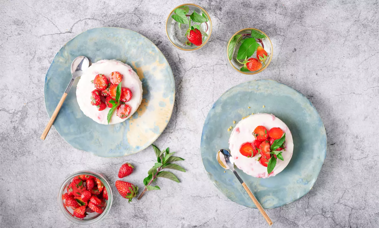 Мини чийзкейкове с бяла, кремообразна повърхност, гарнирани с яркочервени ягоди и свежи листа мента върху небесносини чинии