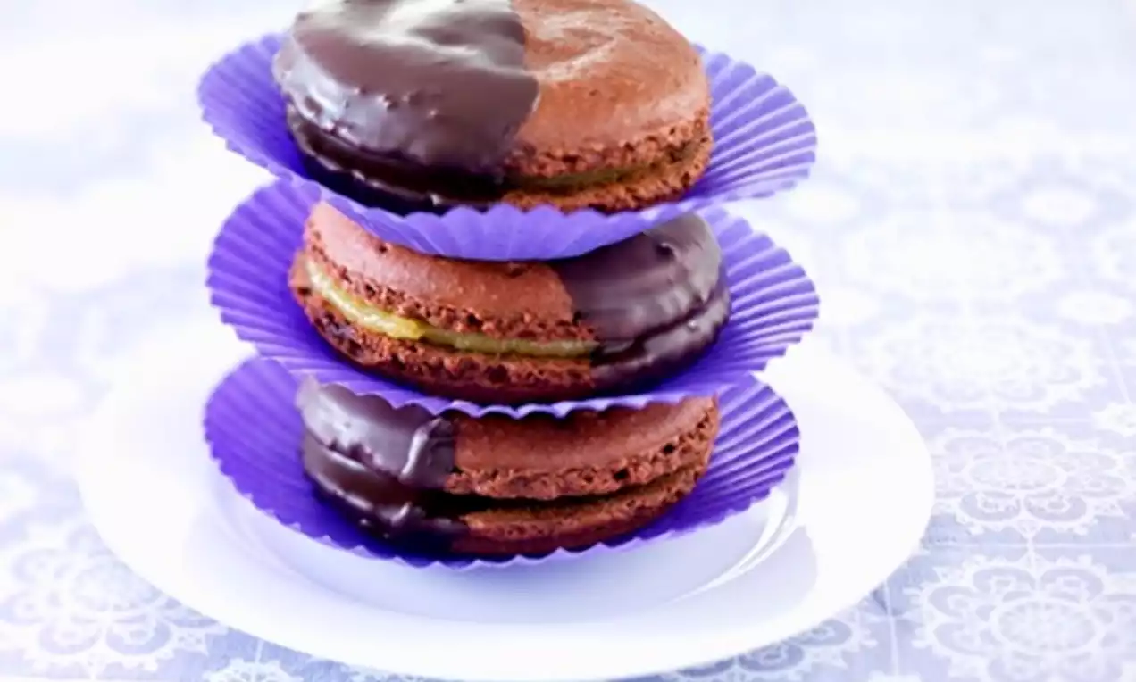 Bruine chocolademacarons met glanzende chocoladehelften gestapeld in paarse papieren vormpjes op een wit bord tegen een kanten tafelkleed
