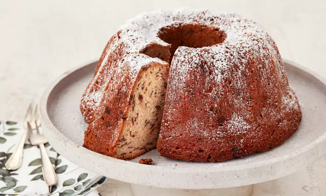 Goldbrauner Gugelhupf mit feuchter Struktur und dunklen Bananenstücken, mit Puderzucker bestäubt auf einer hellen Kuchenplatte angerichtet