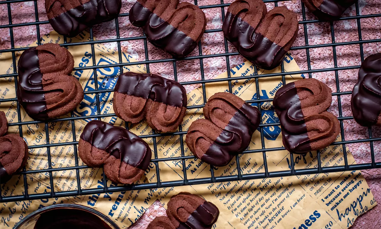 Какаови бисквити с извита форма, потопени наполовина в лъскава шоколадова глазура, подредени върху метална решетка и хартиена подложка