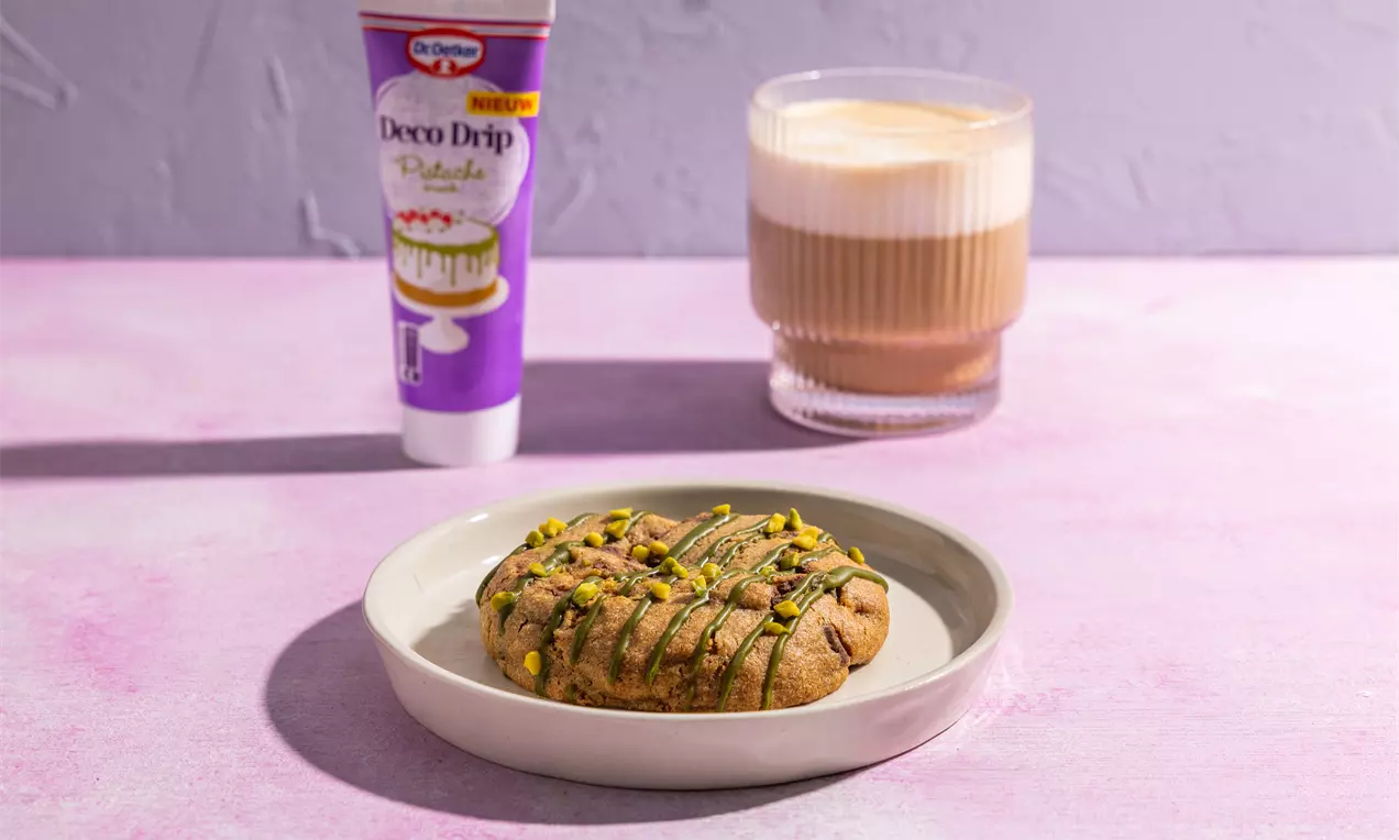 Ronde pistachekoekjes met een goudbruine kleur, groene stukjes pistache en dunne lijnen donkere chocolade op een licht bord, met een latte en decoraties in de achtergrond