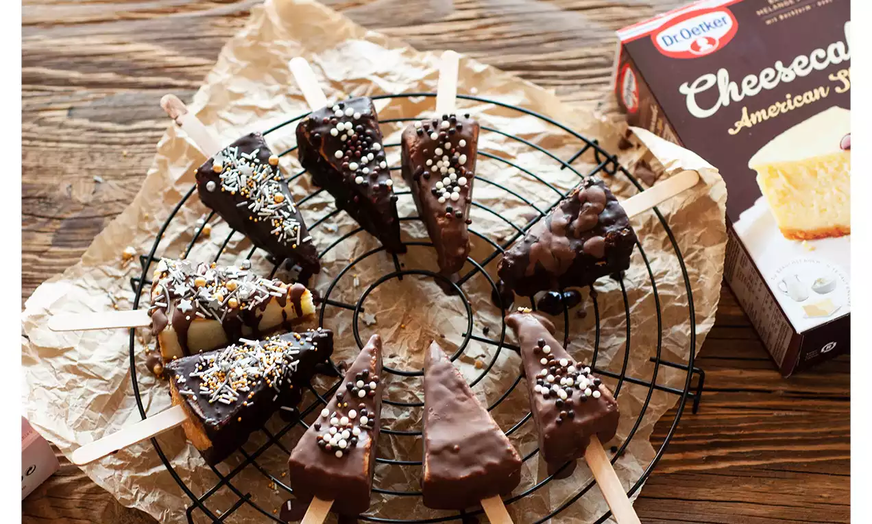 Cheesecake-Sticks in Dreiecksform, mit Schokolade überzogen und bunten Streuseln dekoriert, auf einem schwarzen Kuchengitter angerichtet