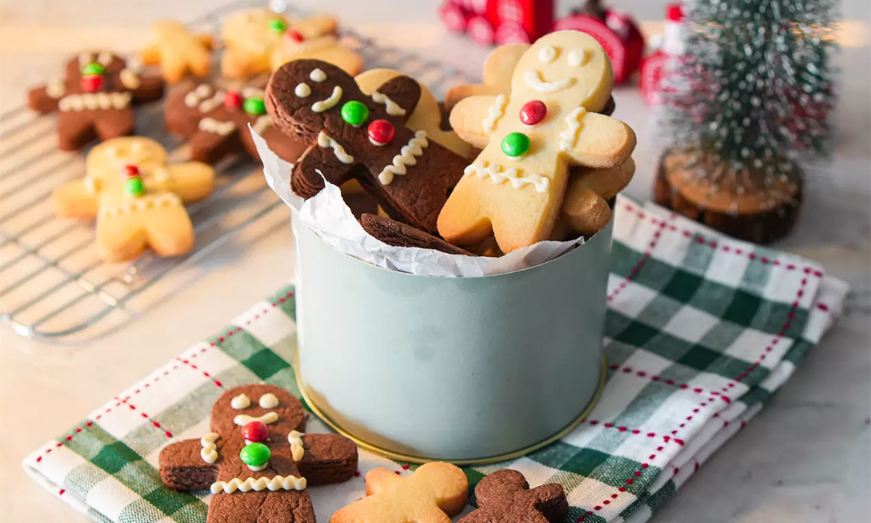 Biscoitos natalinos em forma de bonequinhos, decorados com glacê e confeitos coloridos, dentro de uma lata e espalhados sobre um pano xadrez verde e branco.