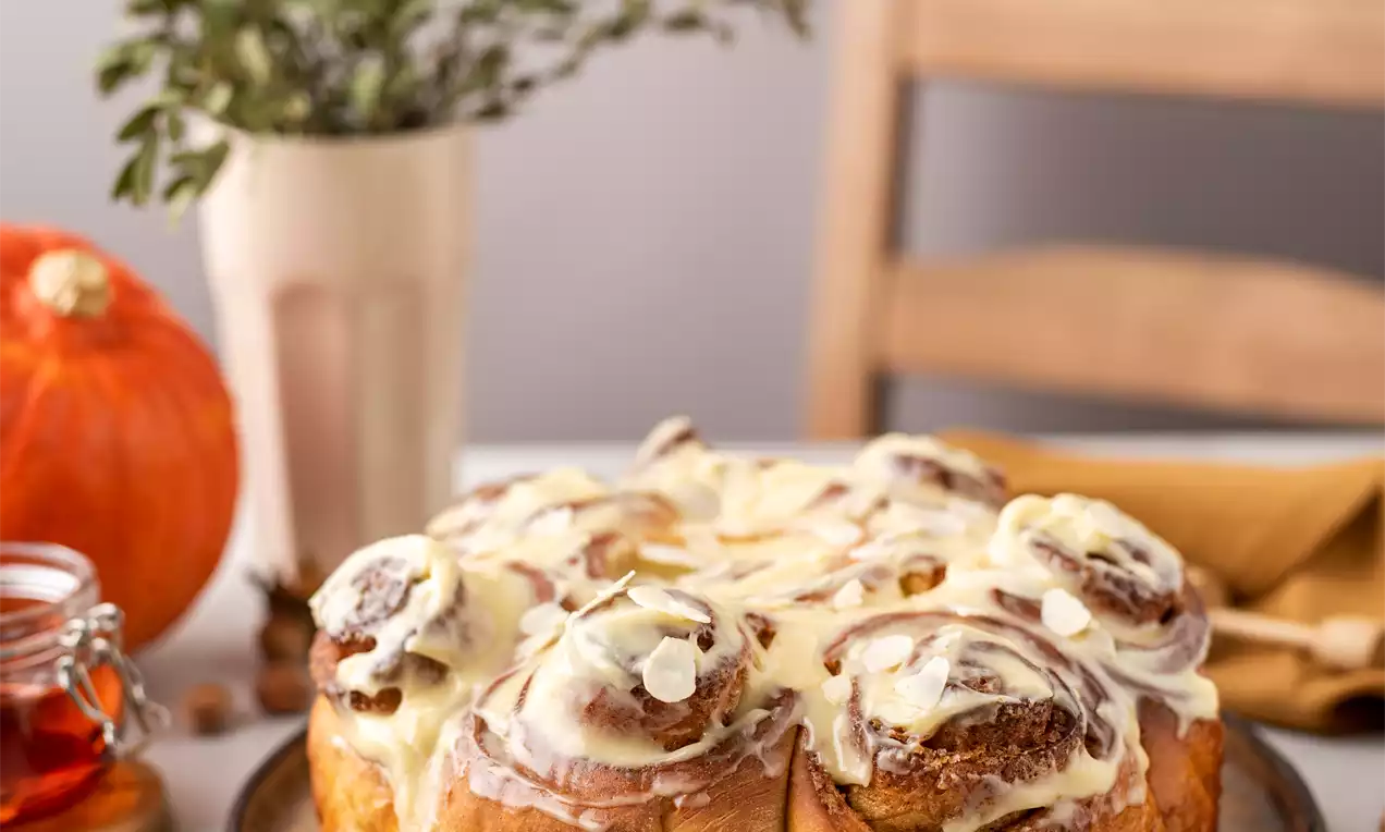 Auksinės purios moliūginės bandelės, susuktos į spiralės formą, gardintos glaisto sūkuriu, patiektos ant apvalios metalinės lėkštės, šalia rudeniškų dekoracijų