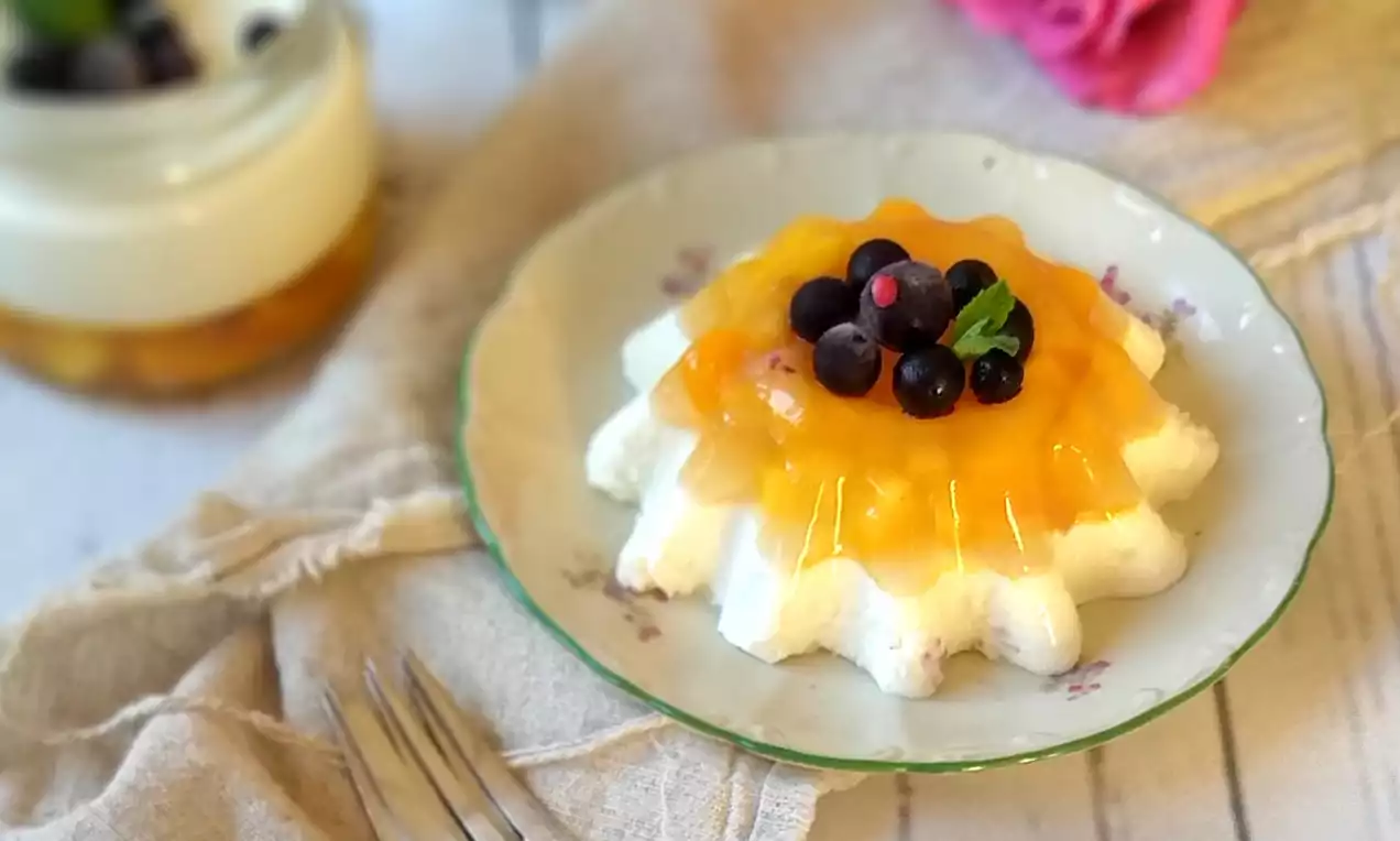 Panna cotta ve tvaru vějířovitého květu s jemnou bílou texturou a hladkým mangovým želé, zdobená lesklými bobulemi na květovaném talíři