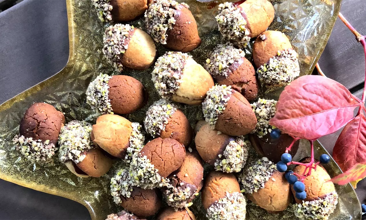 Малки сладки във форма на жълъди с кафяво-златисти половинки, потопени в шоколад и орехи, подредени върху стъклена чиния-звезда с есенна декорация