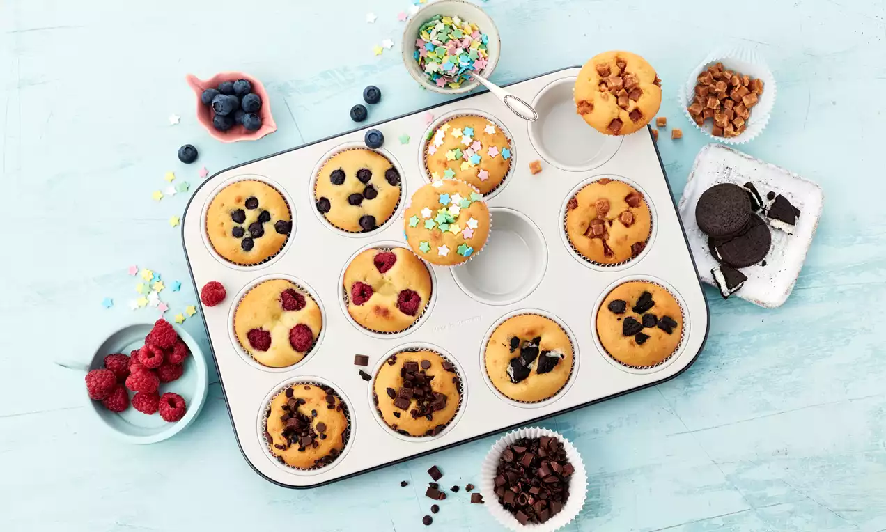 Party-Muffins in einer silbernen Backform, goldgelb gebacken, garniert mit Schokostückchen, Beeren, Streuseln und Seitenbeilagen