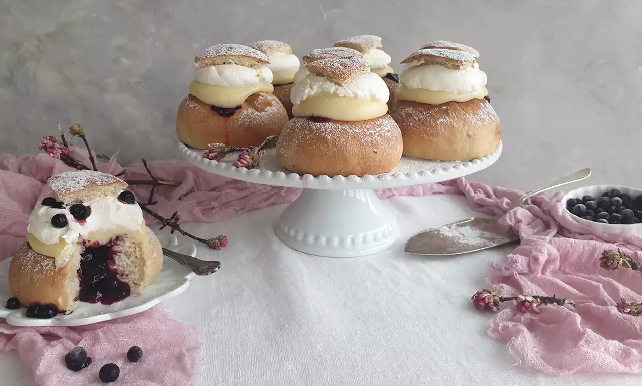 Vaniljsemlor med gyllene bullar, vispad grädde och florsocker, fyllning av vaniljkräm och blåbär på vit kakbricka med rosa tyg och bärskål