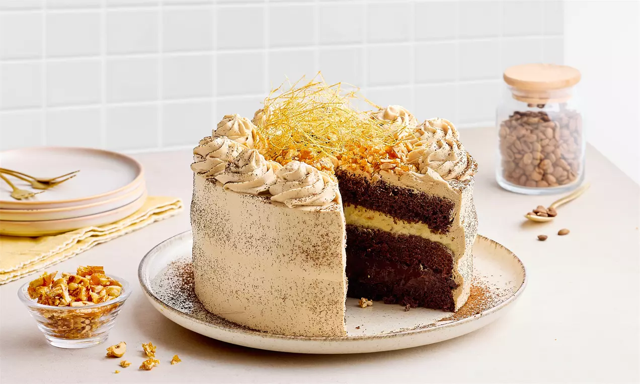 İki katlı kahverengi kek katmanları arasında kremalı dolgu, yüzeyde kahve kreması süslemeleri ve ince krokan parçaları ile tabağa yerleştirilmiş