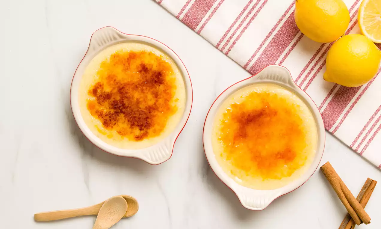 Crema catalana de tonos dorados y caramelo con superficie flameada servida en ramequines blancos junto a limones frescos, canela y cucharas de madera sobre mantel a rayas