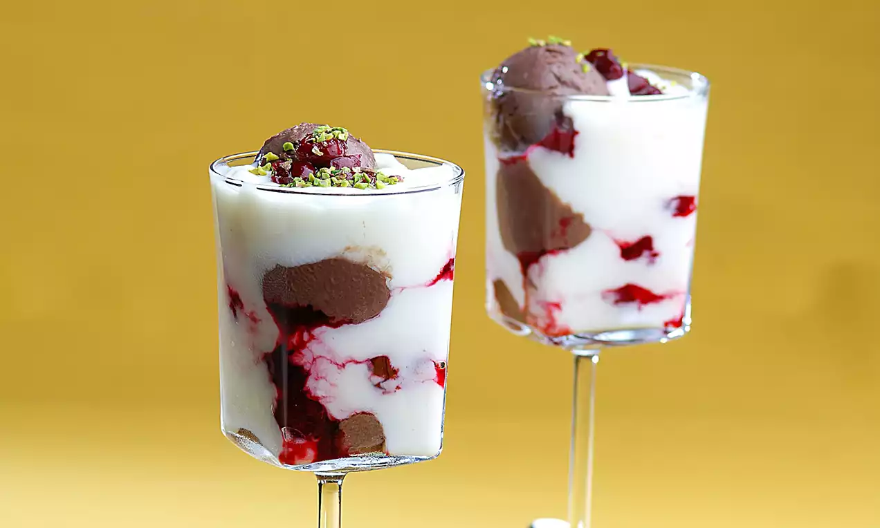 Uzun ayaklı şeffaf bardakta sunulan beyaz sakızlı muhallebi, kırmızı vişne sosu, çikolatalı köpük ve antep fıstığı parçalarıyla süslenmiş