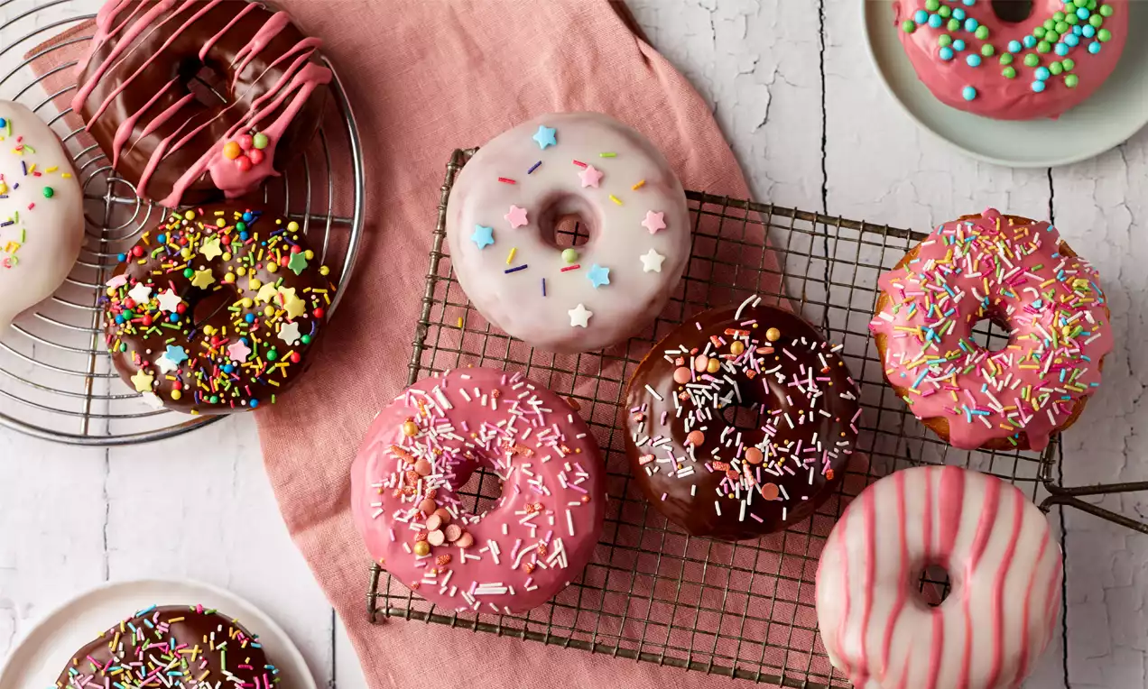 Bunte Donuts mit glänzender Glasur, Zuckerstreuseln und Sternchen, teils auf einem Gitter und Tellern auf rosafarbenem Stoff arrangiert