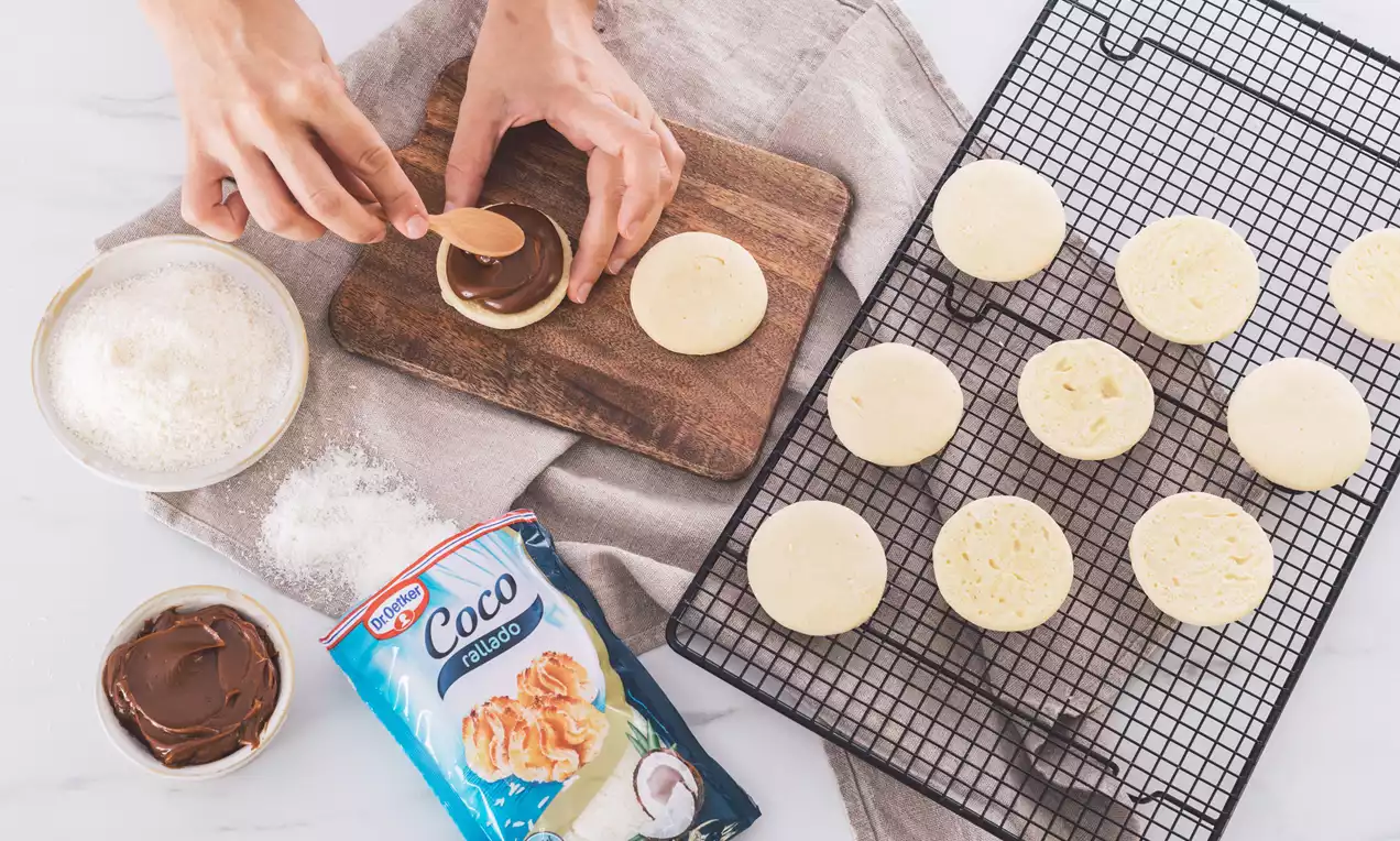 Discos dorados y lisos de masa rellenos de dulce de leche cremoso, bordeados con coco rallado blanco sobre una mesa clara
