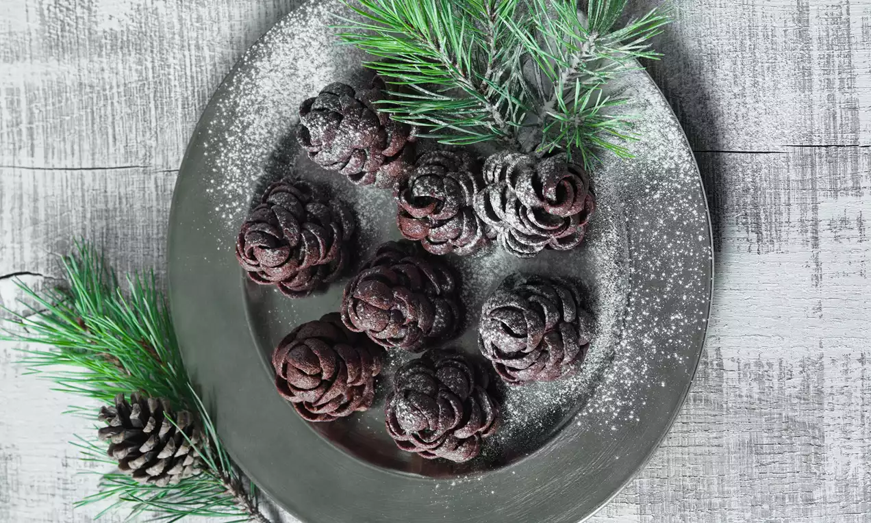 Tmavě hnědé šišky z jemného těsta s vrstvou moučkového cukru na kovovém talíři, obklopeném zelenými větvičkami jehličnanu