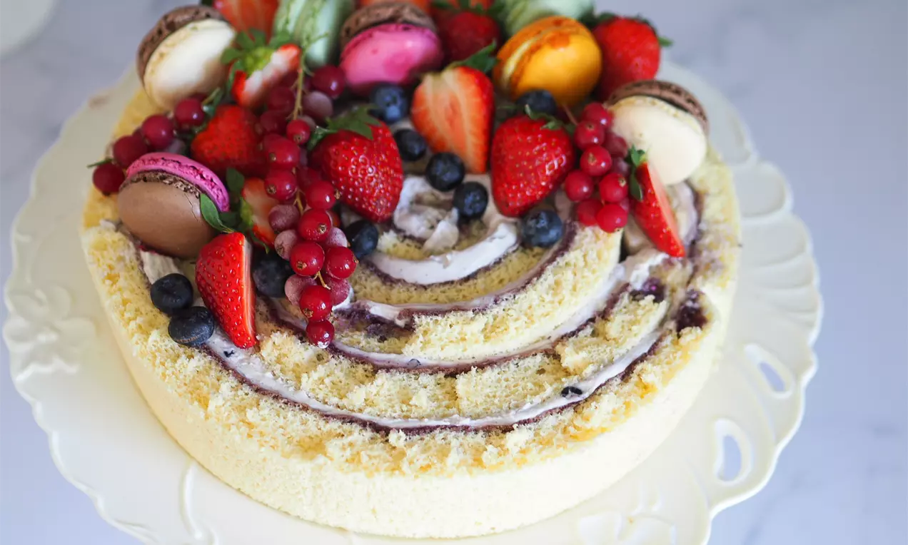 Кръгла вита торта от светъл блат със спираловидно разпределен боровинков крем, украсена с разноцветни макарони, ягоди и касис върху порцеланова чиния