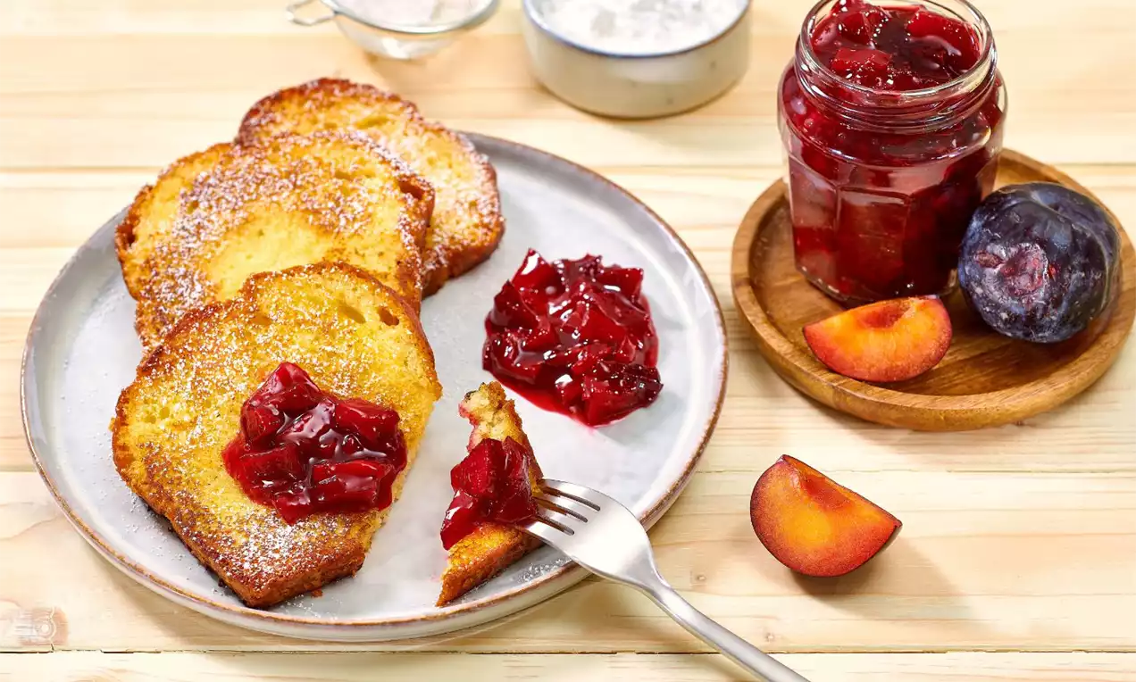 Felii aurii de french toast pufoase, presărate cu zahăr pudră, servite pe o farfurie cu dulceață roșie și prune proaspete pe lemn deschis