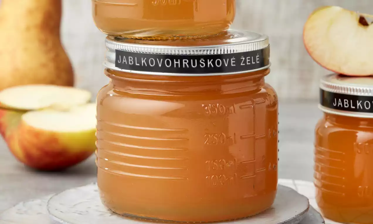 Jablkovohruškové želé jemně oranžové barvy v hladkém skleněném zavařovacím obalu s kovovým víčkem na talíři vedle jablek a hrušek