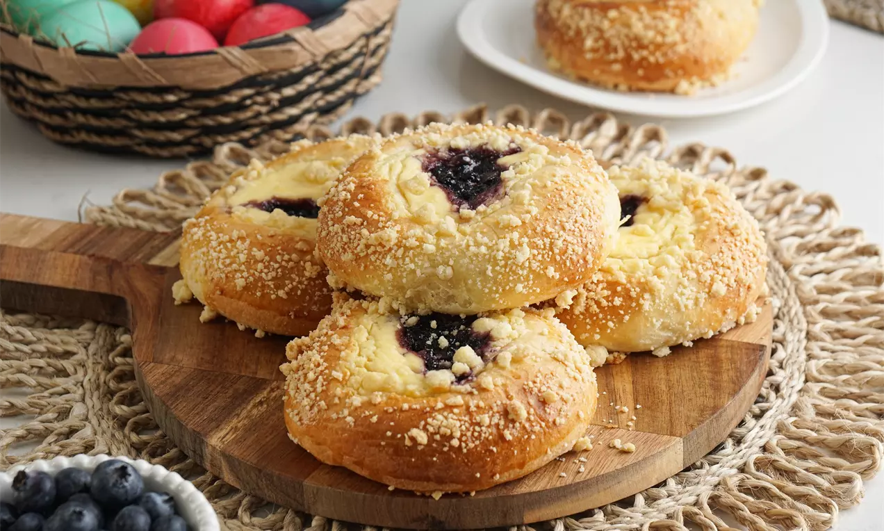 Златисти козуначени питки с ронлива текстура, гарнирани с кремообразен чийзкейк крем и тъмносини боровинки, поднесени върху дървена дъска