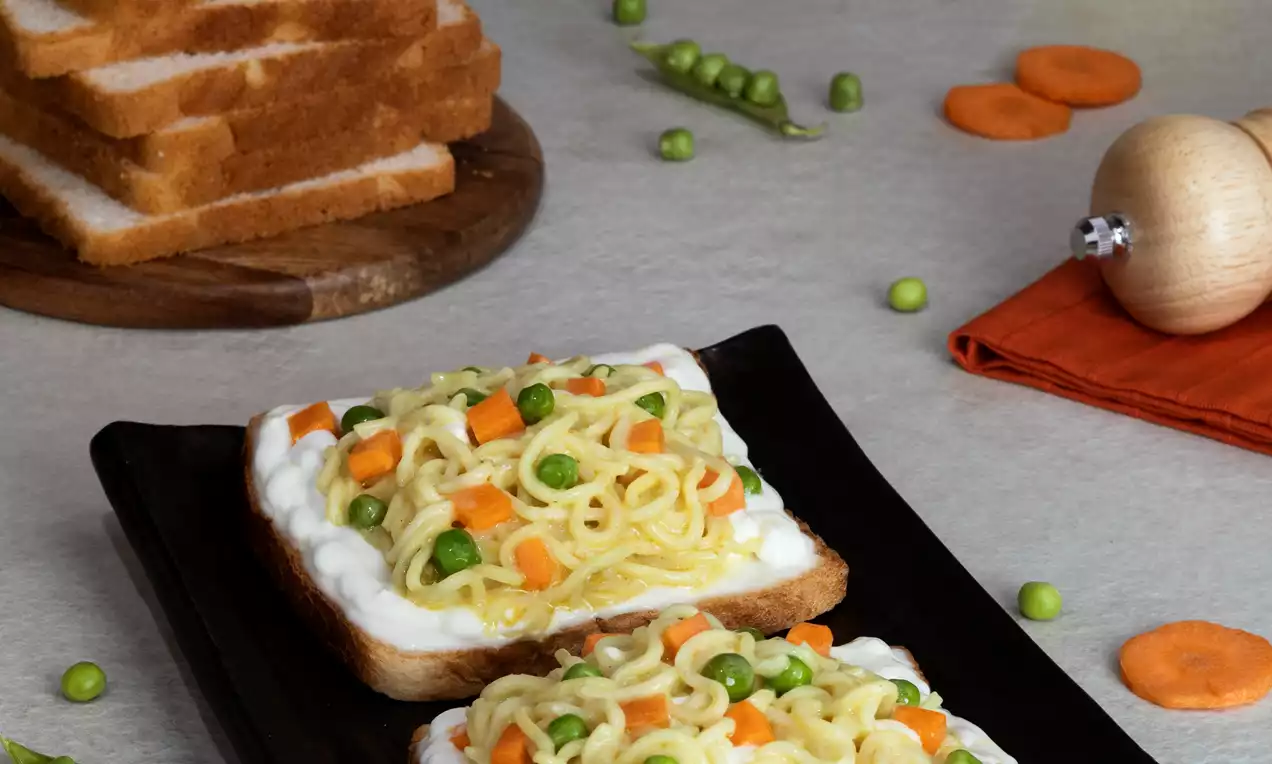 Two slices of toasted white bread topped with creamy white spread, curly yellow noodles, green peas, and orange carrot cubes on a black platter