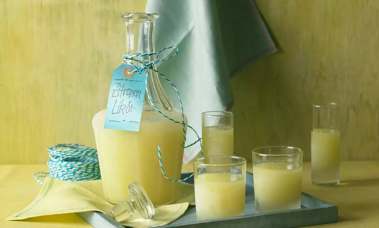Hellgelber Zitronen-Likör in einer bauchigen Glasflasche mit blauem Etikett, serviert in Gläsern auf einem Tablett mit Stoffdeko