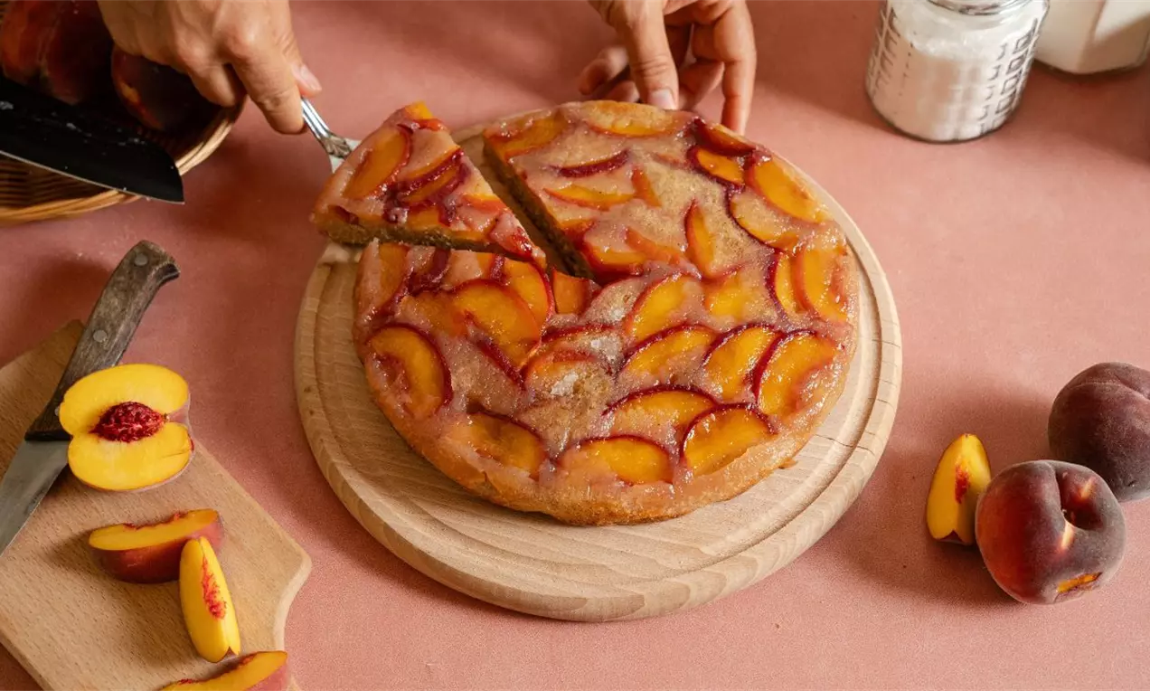 Okrugla torta prekrivena tankim kriškama breskve, na drvenom poslužavniku, okružena celim i sečenim breskvama na stolu boje breskve
