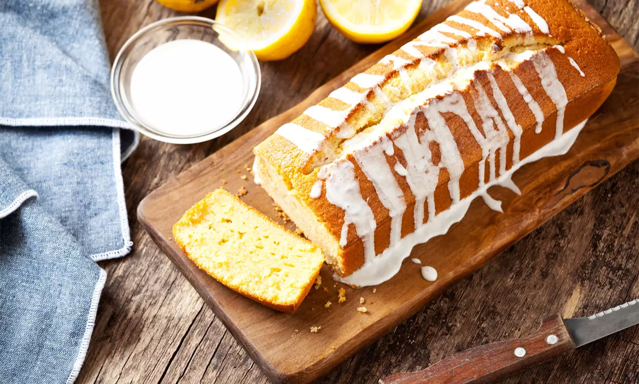 Goudgele rechthoekige citroencake met witte glazuurstrepen op een houten plank, geserveerd naast een mes, halve citroenen en een schaaltje suiker