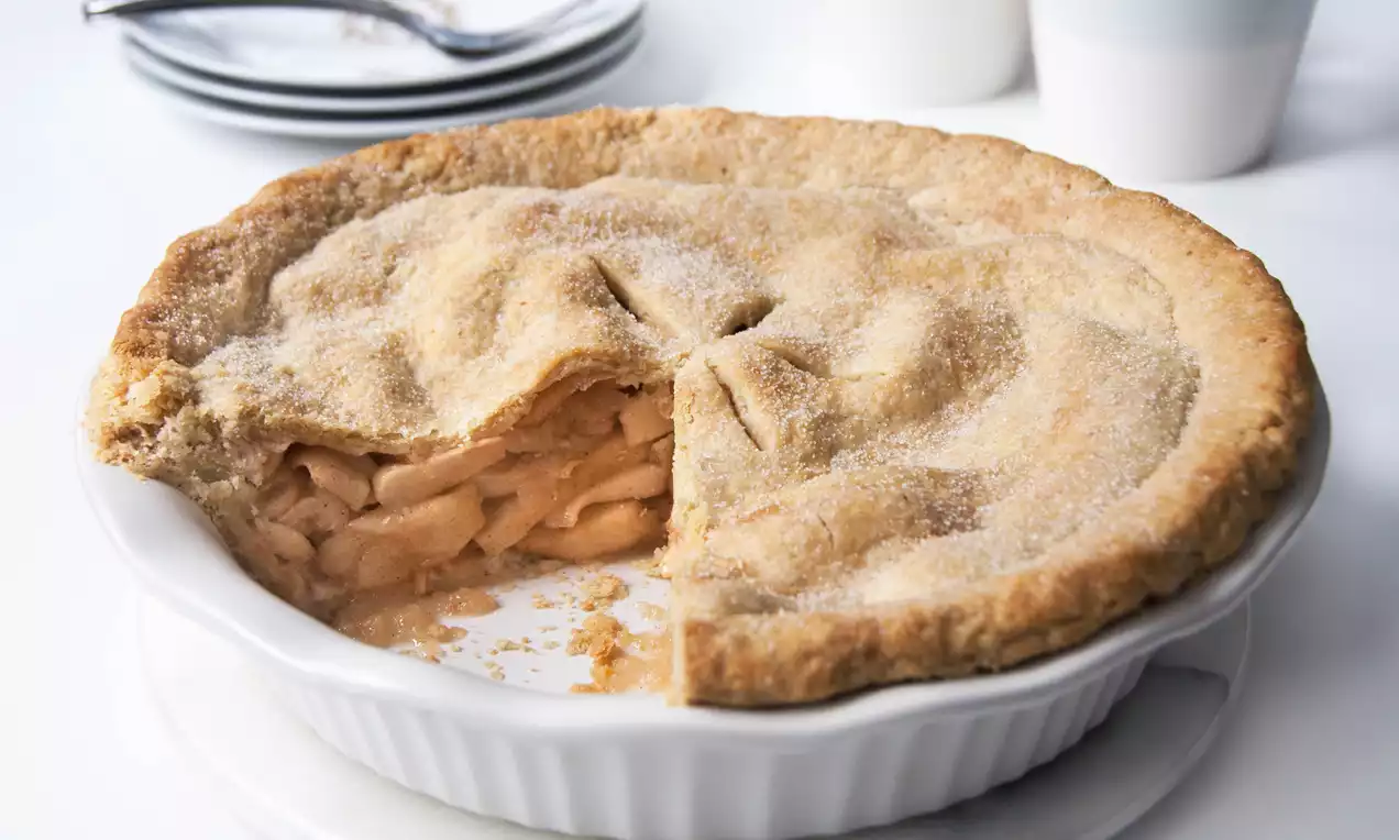 Golden brown apple pie with a sugared flaky crust, a slice cut to reveal tender apple slices inside, in a white ceramic pie dish