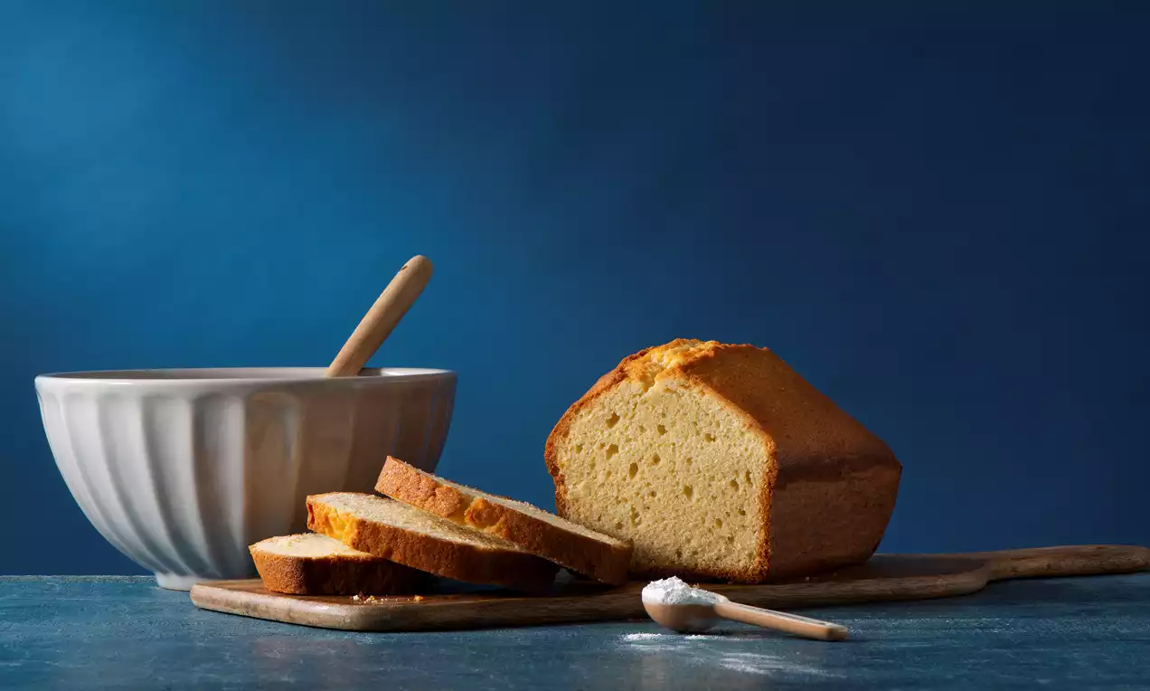 Golden vanilla pound cake with a domed top, soft crumb, sliced on a wooden board next to a white bowl and a small wooden spoon