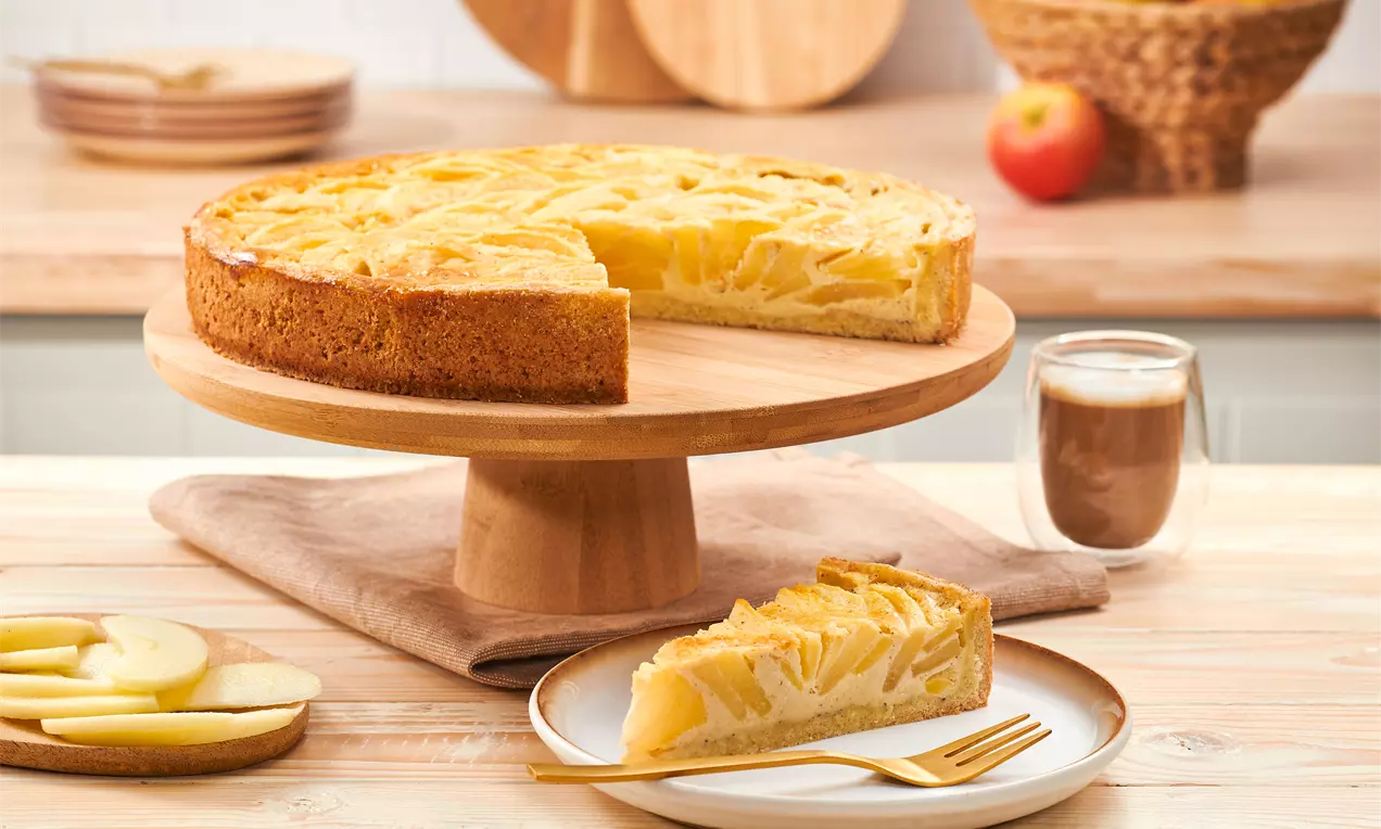 Apfelkuchen mit glänzenden gelben Apfelscheiben und sahnigem Guss auf goldbraunem Boden, serviert auf einer Holzplatte mit Gabel und Apfelscheiben