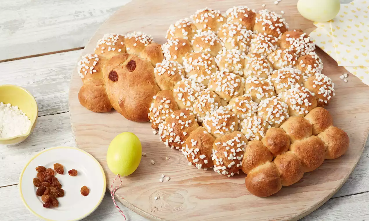 Goldbraunes Hefeteig-Osterlamm mit glänzender Kruste und Zuckerstreuseln, dekorativ auf einem runden Holzbrett neben Ostereiern und Rosinen