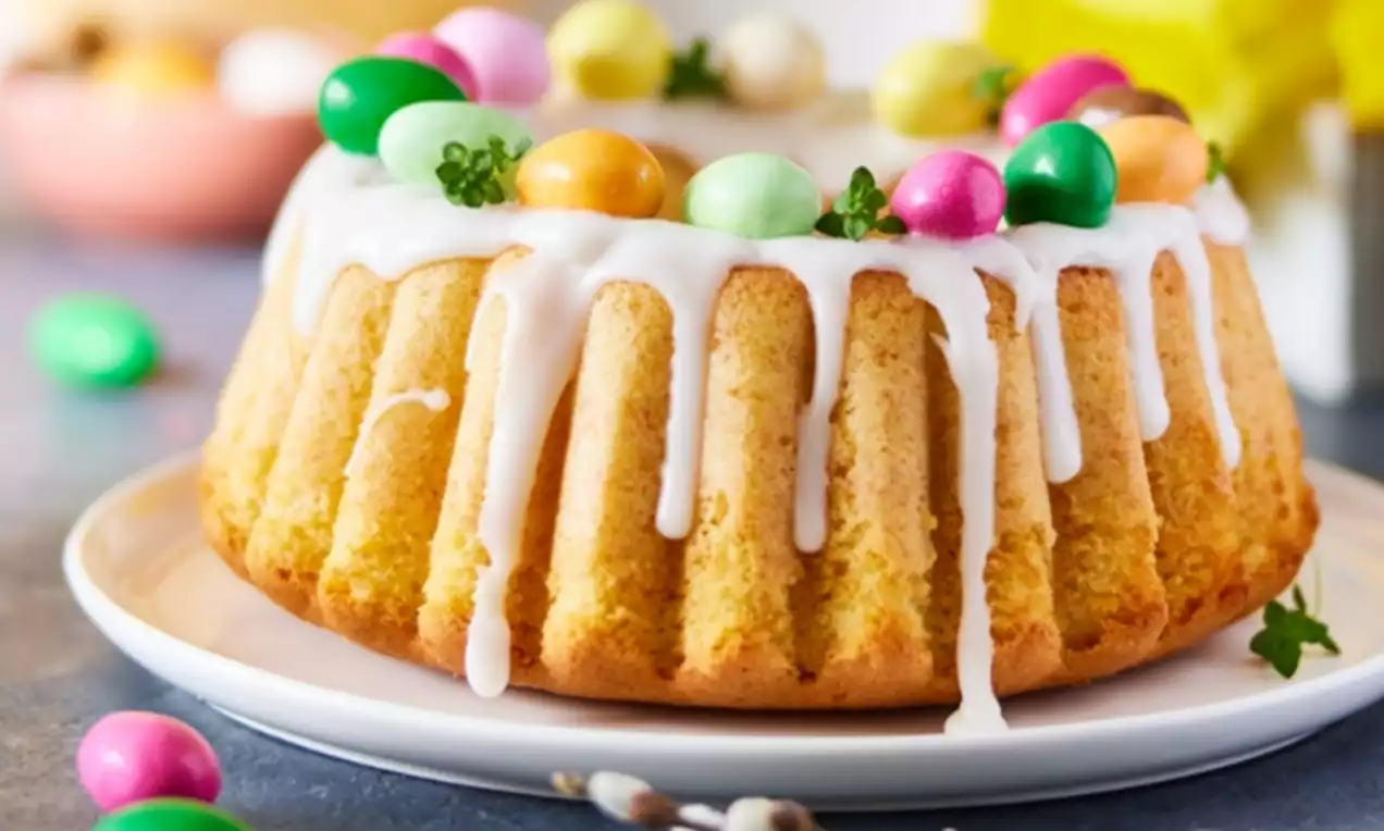 Zeltaini dzeltens kēkss ar baltu glazūru, kas tek pāri maliņām, dekorēts ar košiem olas formas saldumiem un zaļiem augiem uz balta šķīvja