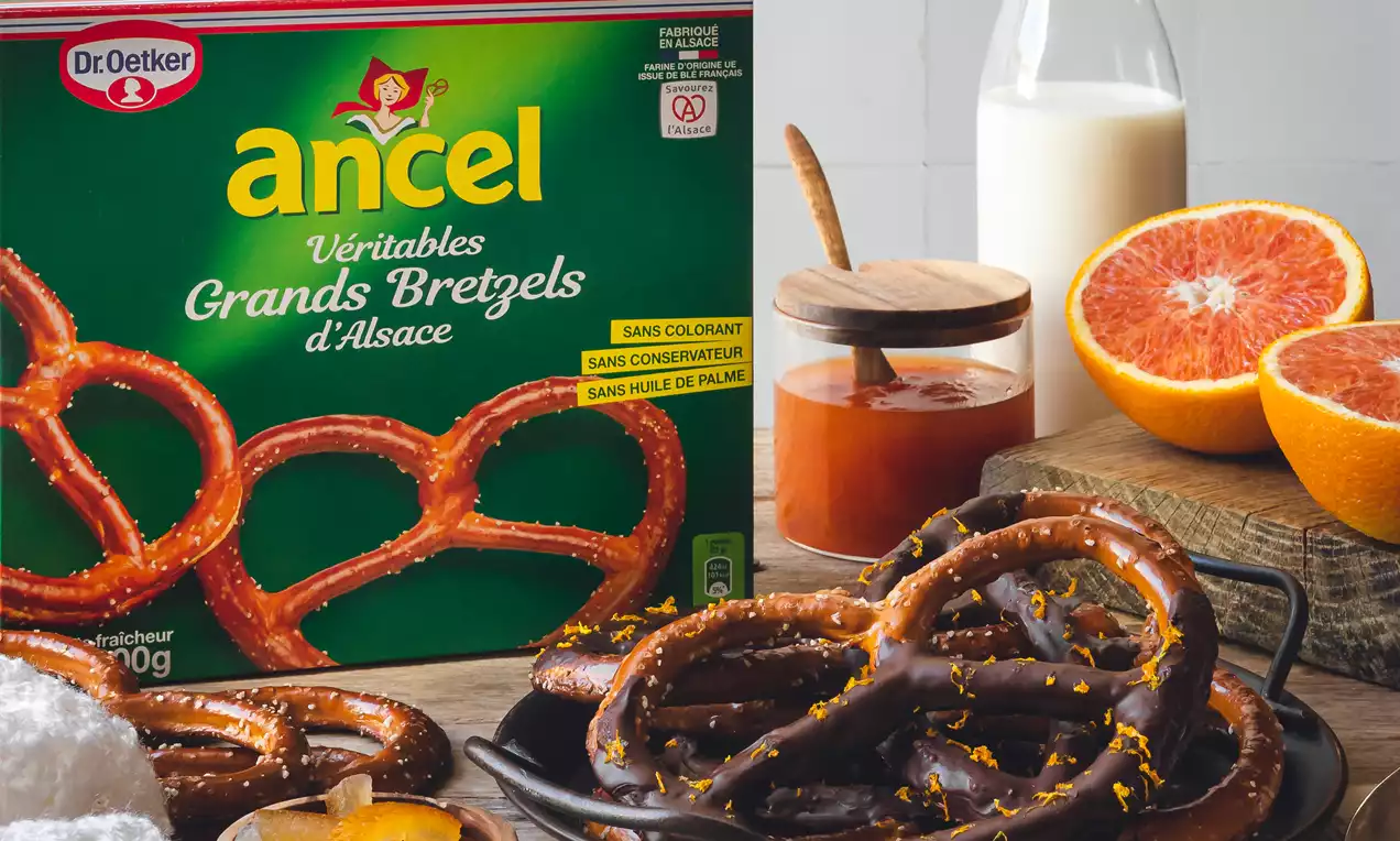 Grands bretzels dorés et brillants avec une texture lisse et brillante, sur une assiette noire accompagnés d'oranges et de confiture