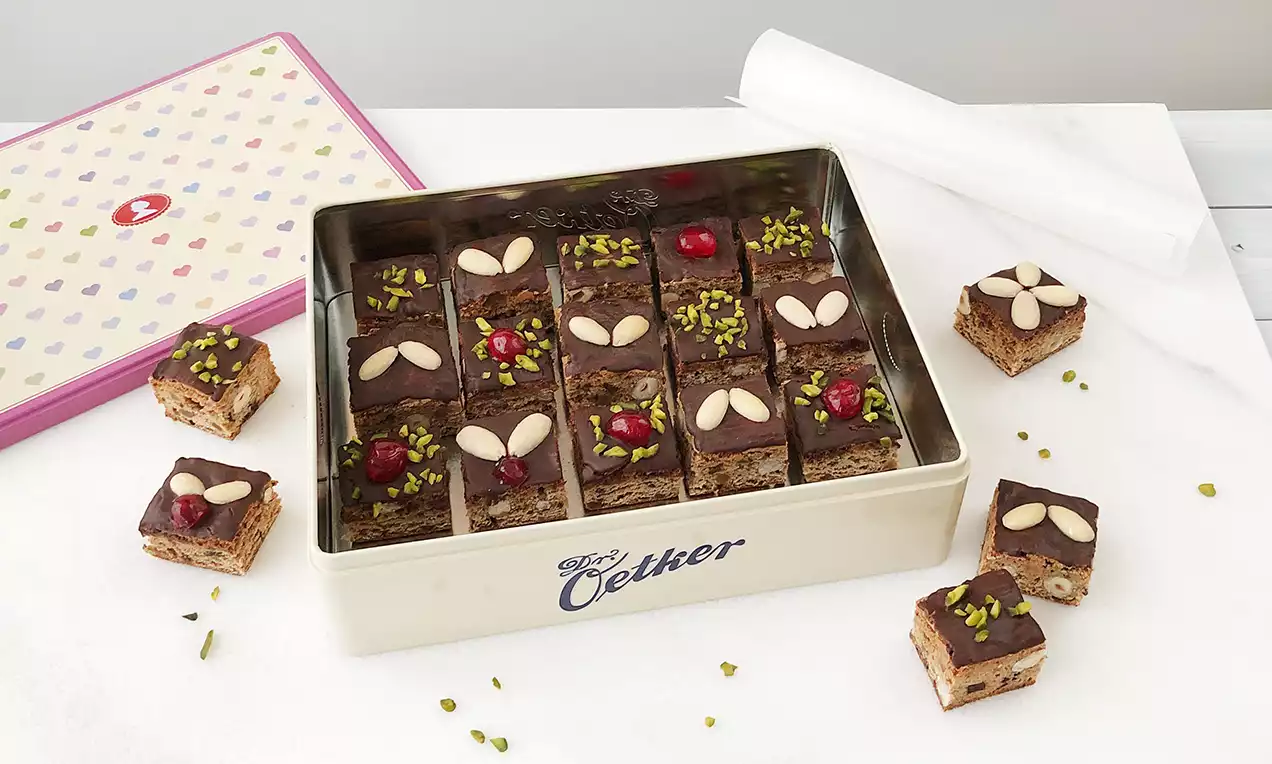 Lebkuchenwürfel mit glänzender Schokoladenglasur, dekoriert mit Mandelsplittern, Pistazien und Kirschen in einer beige-roten Metallbox