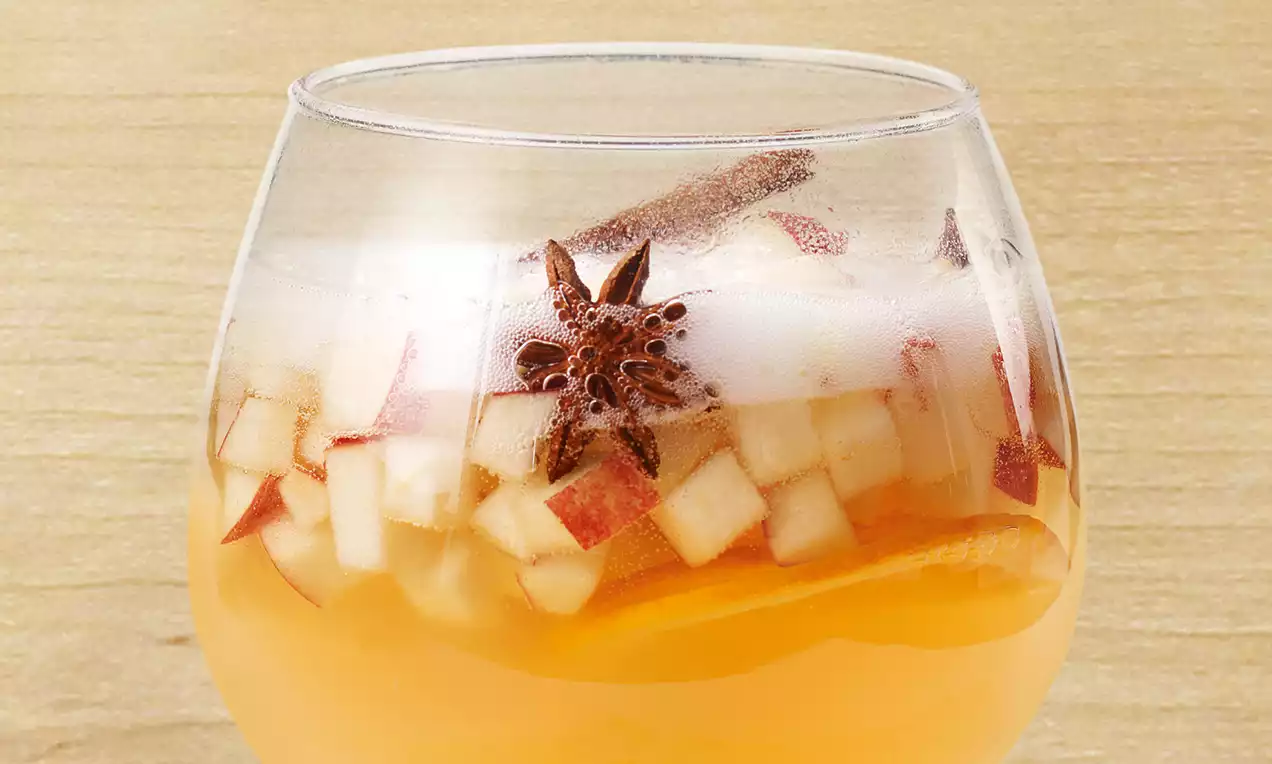 Punsch ambré dans un verre rond garni de dés de pomme rouges et blancs, anis étoilé et bâton de cannelle sur fond de bois