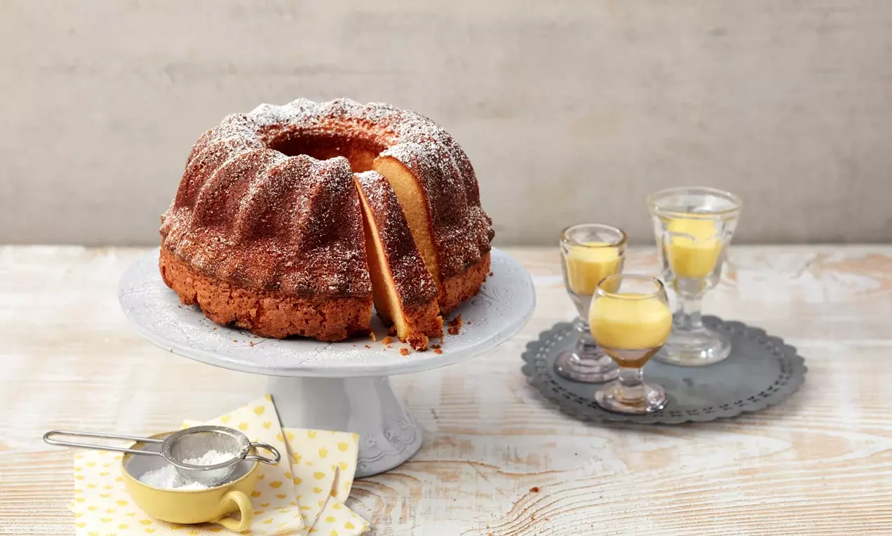 Кекс с яйчен ликьор с златистокафява кора и ронлива текстура, нарязан, поръсен с бяла пудра захар, сервиран върху стойка