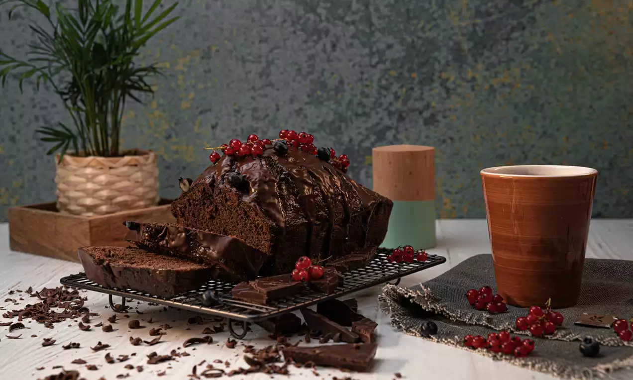 Chec lucios de ciocolată, glazurat și decorat cu coacăze roșii, feliat pe un suport metalic, înconjurat de bucăți de ciocolată și cioburi