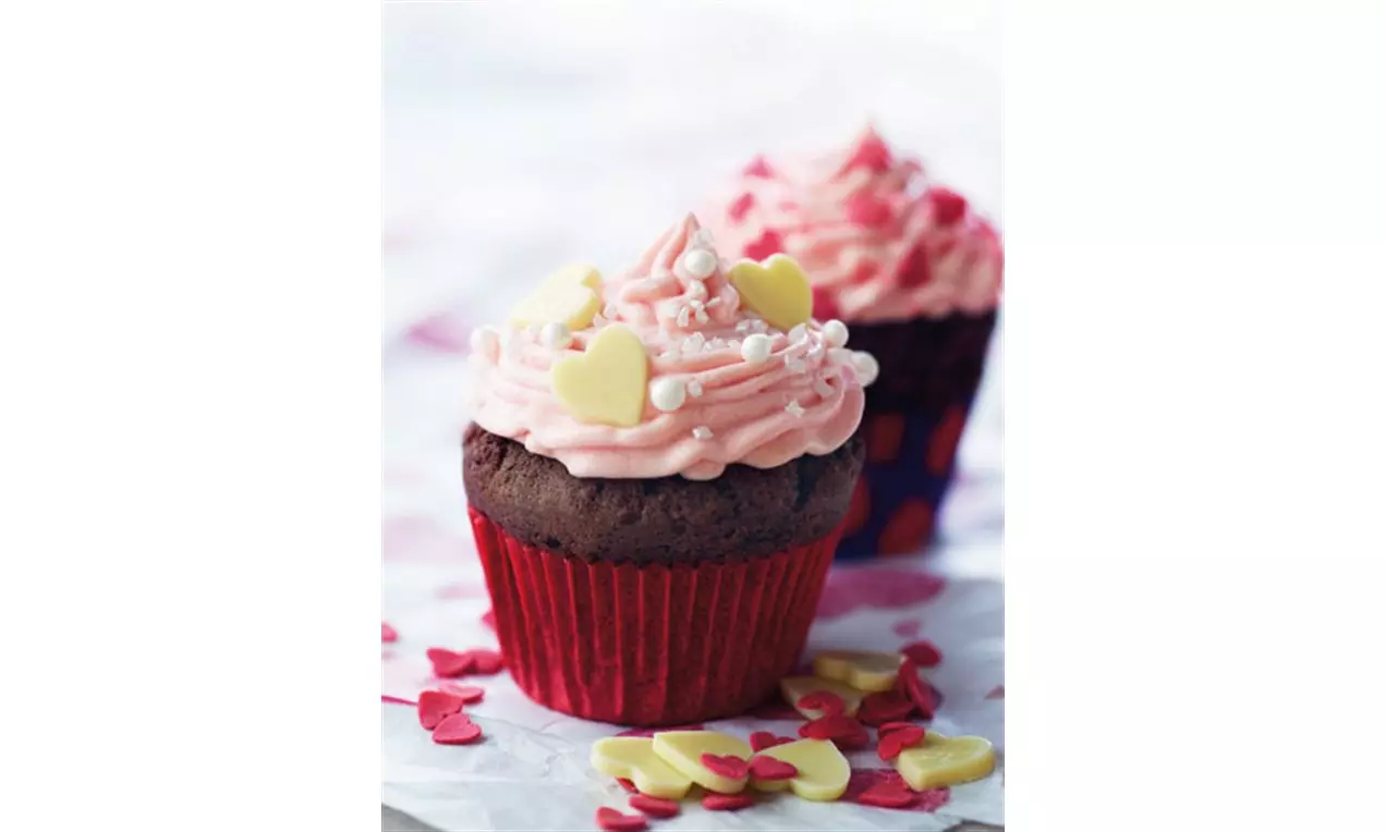 Sjokoladecupcakes i røde former med rosa kremtopp dekorert med hjerteformet gul sjokolade og sukkerstrø på hvitt papir med røde og gule pynt