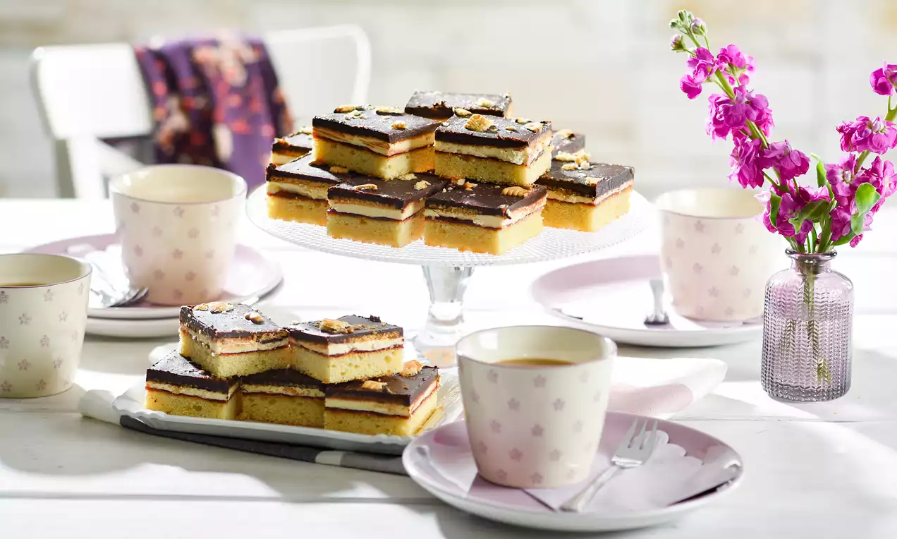 LPG-Kuchen mit goldgelbem Boden, dunkler Schokoladenschicht und Nusskrümeln auf Glasplatte, umgeben von blassrosa Geschirr und Blumenvasen