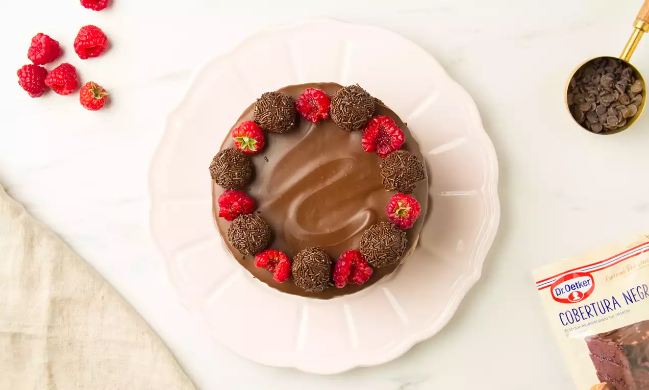 Tarta redonda de chocolate brillante con trufa oscura y frambuesas frescas sobre un plato rosa claro en una mesa blanca