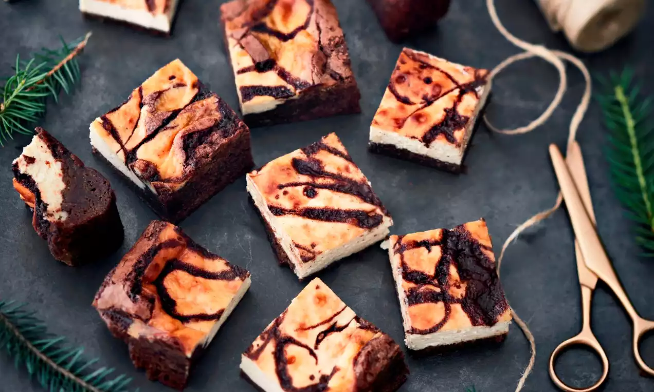 Fyrkantiga brownies med mörk chokladbotten och gyllene mascarponeswirl, placerade på mörk stenbakgrund med grankvistar och guldfärgad sax