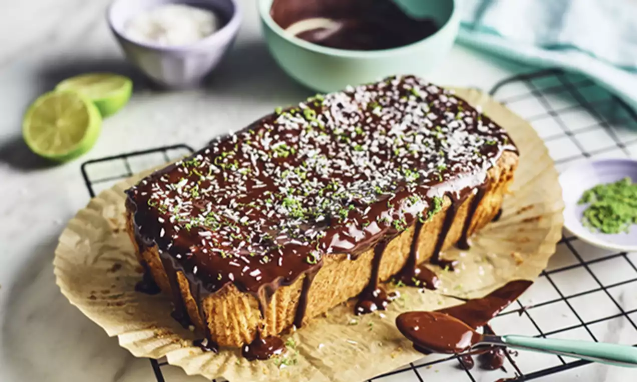 Avlang kokos- og limekake med mørk glasur og dryss av kokos og limeskall på bakepapir, omgitt av limehalvdeler og ulike skåler