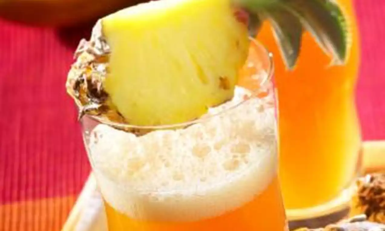 Cocktail aux nuances orange vif avec une mousse légère, décoré d'un morceau d'ananas frais et de son feuillage sur fond coloré