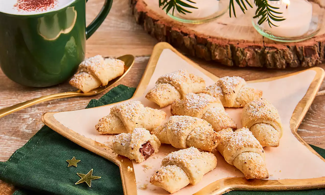 Nugátos kiflik aranyszínű csillag alakú tányéron, enyhén porcukorral szórva, egy kifli tölteléke látszik, mellette zöld terítőn csillagok
