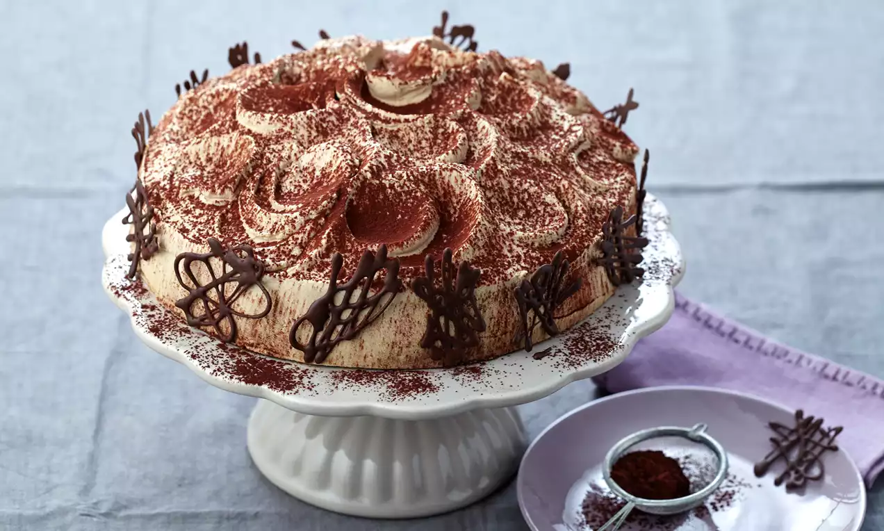 Runde Cappuccino-Sahne-Torte mit geschwungener Creme und Schokodekor auf weißer Tortenplatte, bestäubt mit feinem Kakaopulver