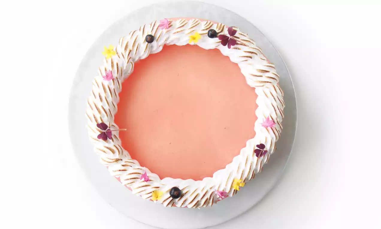 Vegetarisk cheesecake med blank karamel-overflade, hvid marengskrans med gyldne nuancer og små blomster på et hvidt kagefad