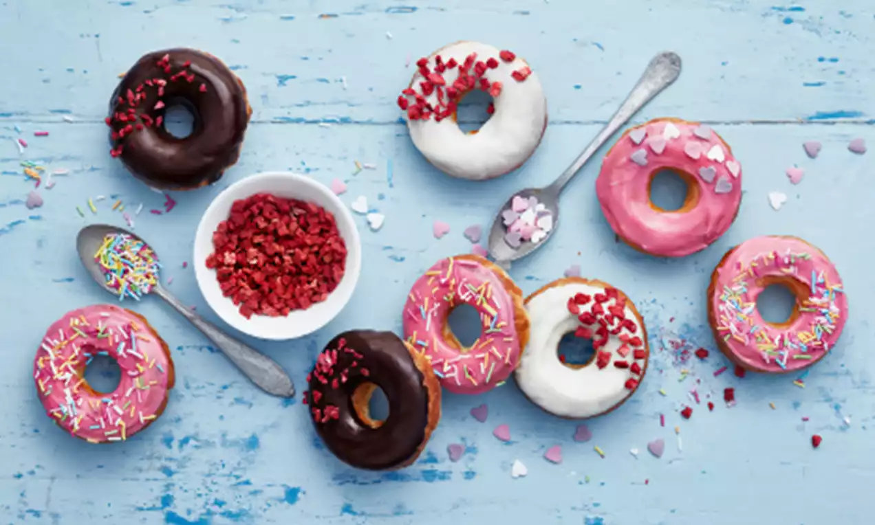 Šarene krafne u boji čokolade, bijele i ružičaste glazure, posute šarenim mrvicama i komadićima voća na svijetloplavoj podlozi