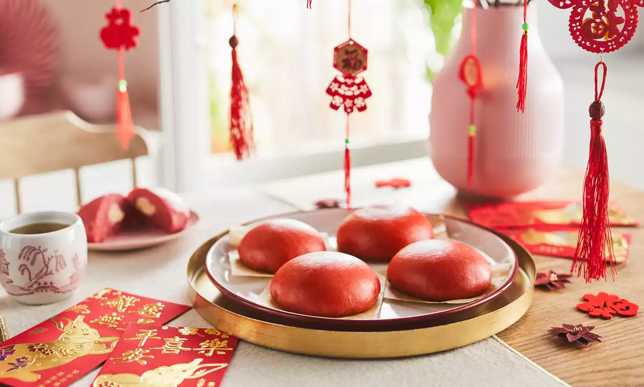 Four smooth, round sweet bao buns with glossy red-orange tops on a plate with festive red decorations and a cup of tea nearby